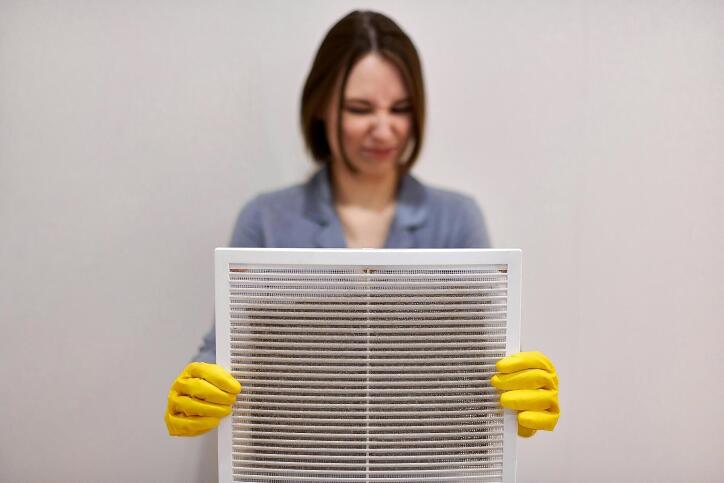 A woman holding a dirty air vent filter with yellow gloves and making a disgusted face at it.